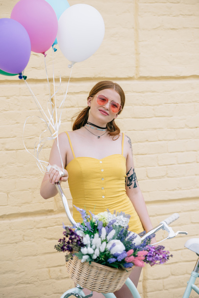 beautiful young woman in sunglasses smiling at camera while standing with bicycle and colorful balloons on street - Photo, Image