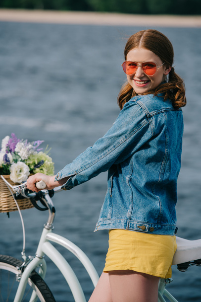 heureux attrayant fille en lunettes de soleil souriant à la caméra tout en se tenant avec vélo près de la rivière
 - Photo, image