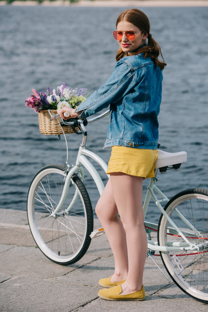 beautiful young woman in sunglasses smiling at camera while standing with bicycle near river - Foto, Imagem
