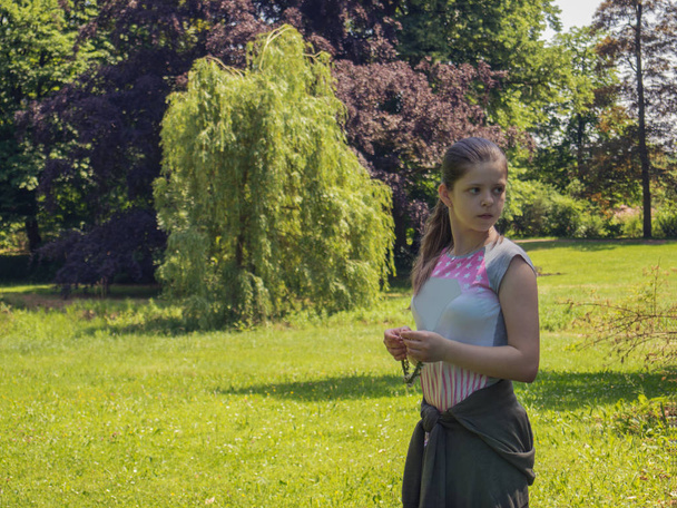 背景の木々 に囲まれた公園の緑の夏の芝生と花と見える、フレーム外の花輪を持ってカメラに半分悲しい孤独な白人の十代女の子はスタンド - 写真・画像
