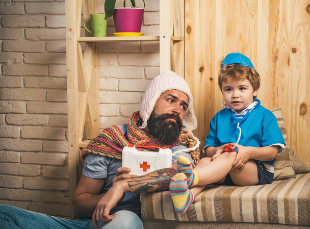 Family time, bearded man with serious boy playing doctor. Dad looks at kid dressed like male nurse with stethoscope. Man in white knitted hat with sad look holding toy first aid box, medicine concept. - Fotó, kép