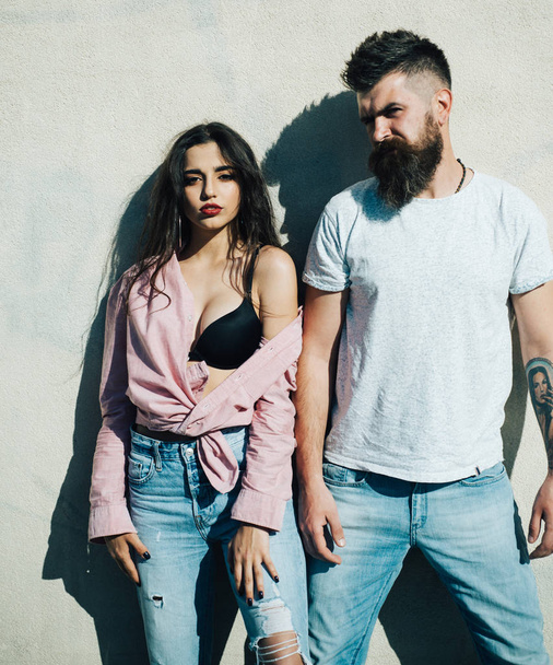 Couple in love by white wall. Brutal bearded man in white T-shirt and brunette girl in boyfriend jeans and pink oversize shirt over black bra with impudent look outdoors, rebellious youth concept. - Фото, изображение