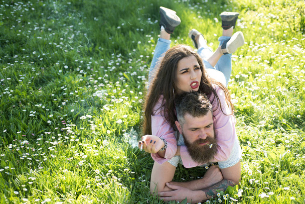 Smoking habit concept. Couple in love spend time outdoors and smoking. Man and woman laying on grass spring day. Couple on strict faces smoking outdoors, nature on background. - Photo, Image
