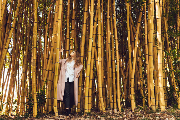 curly young woman in long pink coat posing next to tall bamboo trees, in park - Foto, Imagem