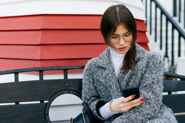 ένα κορίτσι νεαρή μελαχρινή πόλη σε ένα μοντέρνο γκρι παλτό και γυαλιά κάθεται σε έναν υπαίθριο πάγκο και γράφει ένα μήνυμα στο smartphone σε - Φωτογραφία, εικόνα