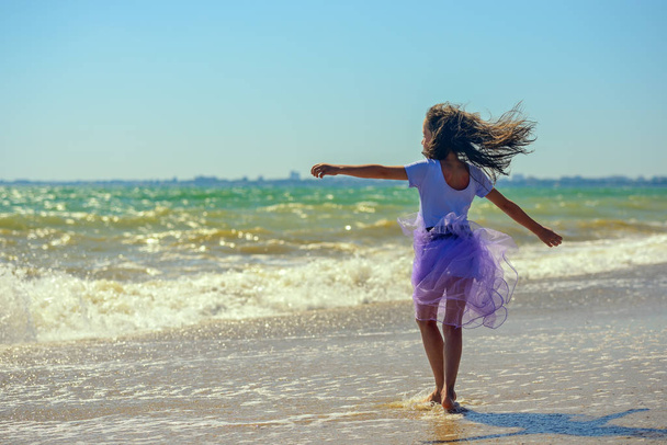Happy little girl is spinning and dancing on the beach on a Sunny day in a purple dress against the background of raging waves. Her hair is fluttering in the wind. The horizontal orientation. - Foto, afbeelding