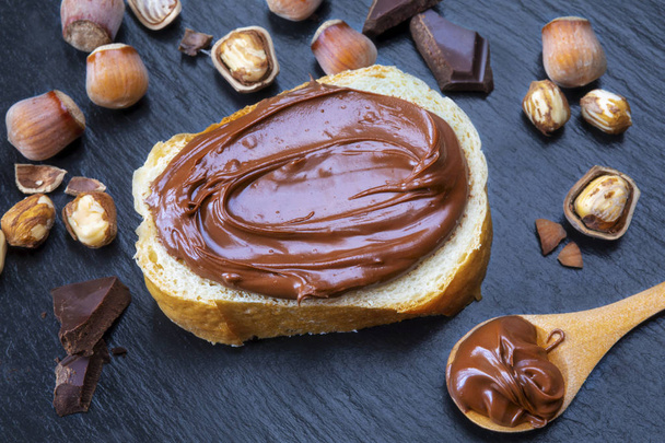 Slice of bread with spread chocolate cream and hazelnuts on black table. Serving a delicious breakfast concept. - Photo, Image