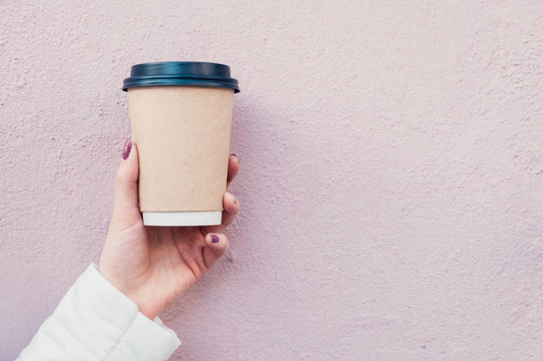 hand holds coffee in brown cardboard cup with black cap on pink wall background - Foto, imagen