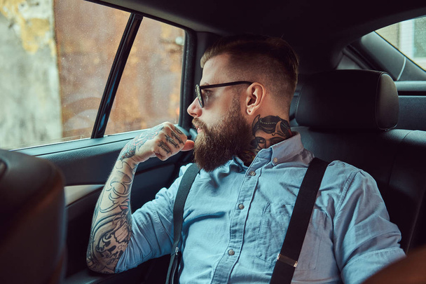 Pensive old-fashioned tattooed hipster guy in a shirt with suspenders sitting in a luxury car on a back seat, looking away. - Photo, Image