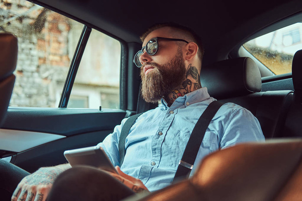 Ο τύπος ντεμοντέ τατουάζ hipster σε ένα πουκάμισο με ζαρτιέρες που κάθεται σε ένα πολυτελές αυτοκίνητο στο πίσω κάθισμα. - Φωτογραφία, εικόνα