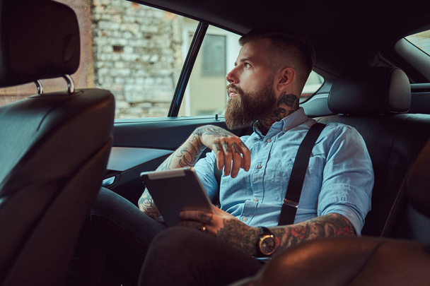 Τύπος της ντεμοντέ τατουάζ hipster σε ένα πουκάμισο με ζαρτιέρες, κρατώντας ένα δισκίο, ενώ κάθεται σε ένα πολυτελές αυτοκίνητο στο πίσω κάθισμα, κοιτάζοντας μακριά. - Φωτογραφία, εικόνα