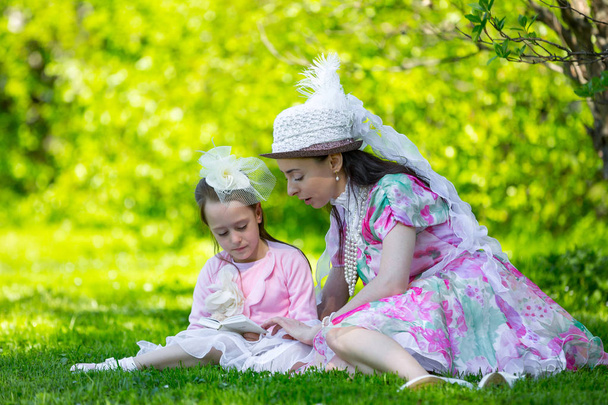 Family idyll, sitting on green grass in a sunny bright park in beautiful light vintage dresses in beads and hats, reading books and fooling around together. Mother and daughter in a summer park. - Foto, Imagen