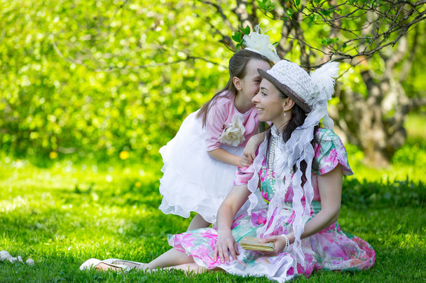 A little girl hurries to share secrets with her mom in the ear in a bright sunny park on the green field in beautiful white classic aristocratic dresses. Mother and daughter in a summer park. - Фото, изображение