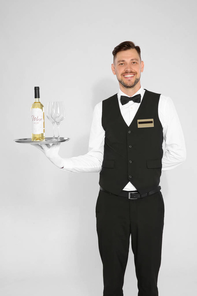 Handsome waiter holding tray with glasses and bottle of wine on light background - Photo, Image