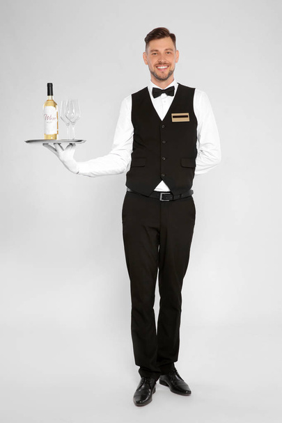 Handsome waiter holding tray with glasses and bottle of wine on light background - Foto, Imagen