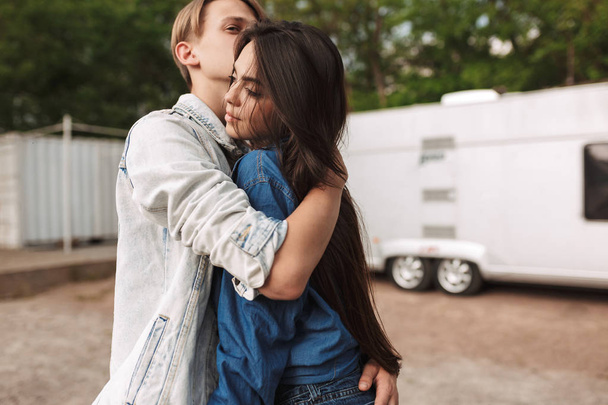 Retrato de una hermosa pareja joven de pie y abrazándose mientras pasan tiempo juntos en el parque con remolque gris en el fondo
 - Foto, Imagen