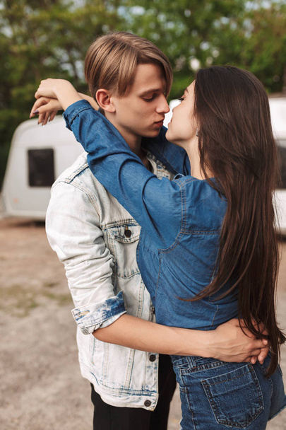 Πορτρέτο της δροσερό ζευγάρι αγκαλιάζει και φιλιούνται ενώ στέκεται στο πάρκο με γκρι ρυμουλκούμενο σε φόντο. Νεαρό ζευγάρι σε μια ημερομηνία - Φωτογραφία, εικόνα