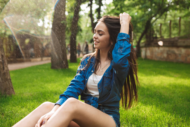 Giovane bella signora felice in pantaloncini e camicia di jeans sogno chiudendo gli occhi mentre si siede sull'erba sotto la pioggia nel parco
 - Foto, immagini