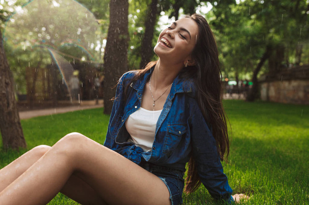 Ritratto di bella signora sorridente in pantaloncini corti e camicia di jeans che chiude gli occhi mentre si siede sull'erba sotto la pioggia nel parco
 - Foto, immagini