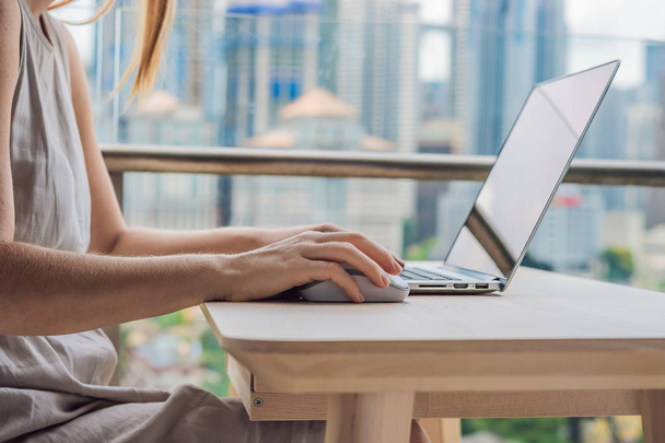 Young woman is working on a laptop on her balcony overlooking the skyscrapers. Freelancer, remote work, work from home - Fotografie, Obrázek