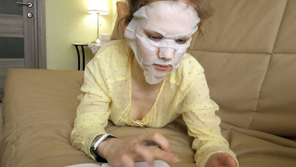 Jeune femme faisant masque facial masque avec masque nettoyant, clique sur le canapé avec smartphone à la maison
 - Photo, image