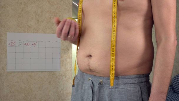 Lihava mies mittaa vyötäröään, isoa kaljavatsaansa, terveellistä elämäntapaansa syöden omenaa.
 - Valokuva, kuva