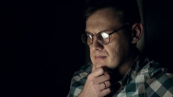 Ένας άνδρας με γυαλιά που λειτουργεί αργά τη νύχτα. Αυτός φαίνεται ανήσυχος, βλέπει στην οθόνη του υπολογιστή μπροστά του, μια στενή-up - Φωτογραφία, εικόνα