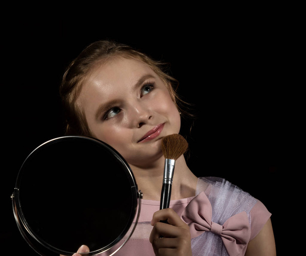 Little girl with round mirror doing makeup on a dark background - Foto, imagen