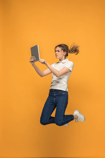 Image of young woman over orange background using laptop computer or tablet gadget while jumping. - Photo, Image