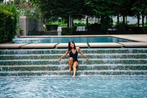A young Chinese Asian woman relaxes in a pool during a hot and sunny day in her bathing suit. She is petite, tanned, toned and muscular and is smiling as she unwinds. - Zdjęcie, obraz