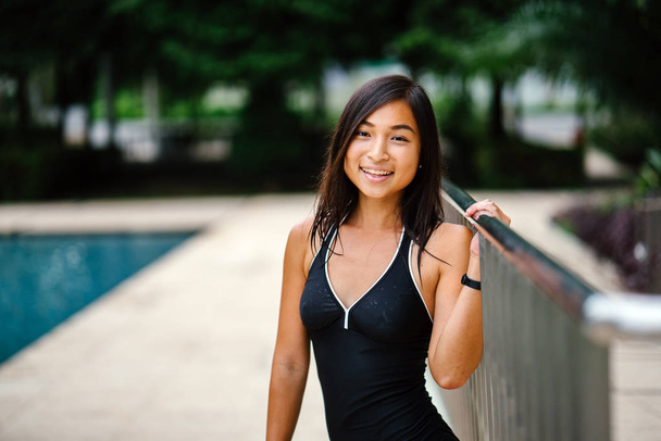 Portrait of a young Chinese Asian girl  by the poolside before her swim on a sunny weekend. She is young, fit, attractive and tanned and is smiling as she takes her shower. - Foto, Imagen