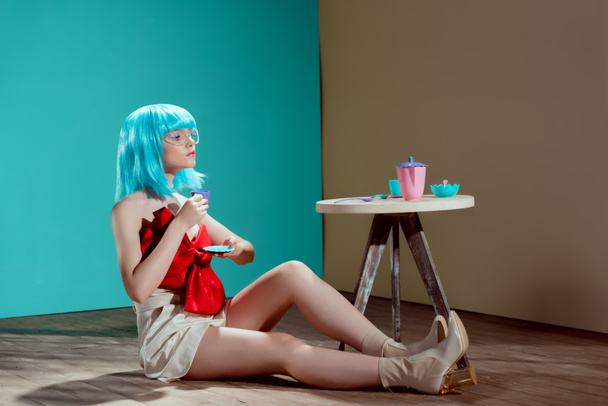 attraente donna elegante in parrucca blu tenendo tazza giocattolo e guardando lontano mentre seduto sul pavimento
   - Foto, immagini