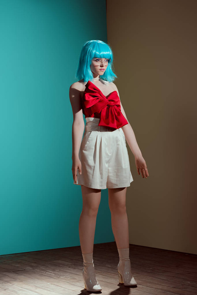 fashionable female model in blue wig and red bow looking away in studio - Фото, зображення