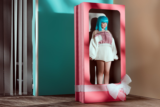 beautiful young woman in fashionable clothes and blue wig posing in decorative box with bow  - Photo, Image