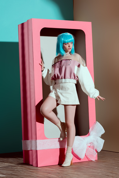 attractive fashionable girl in blue wig posing in decorative pink box with bow and looking away - 写真・画像
