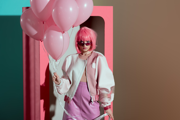 stylish girl in pink wig holding balloons and looking at camera while standing near decorative box  - 写真・画像
