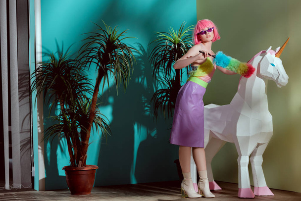 stylowa dziewczyna w różowych peruk gospodarstwa prochowiec i schludny jednorożec stojący - Zdjęcie, obraz