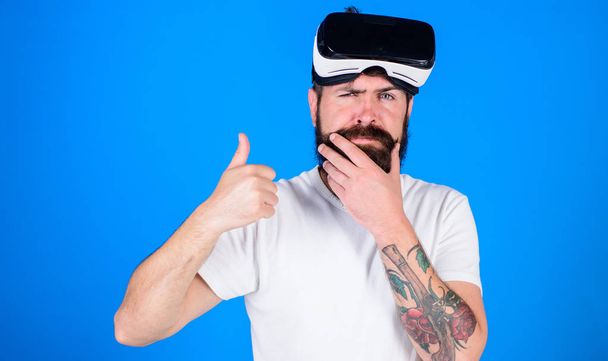 Man with beard and mustache with VR glasses, blue background. VR technology concept. Guy with VR glasses, head mounted display. Hipster on thoughtful face show thumbs up gesture - Фото, изображение