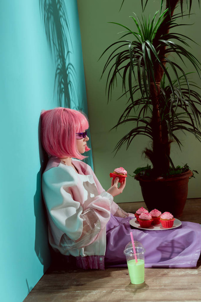 Widok z boku z stylowa dziewczyna w różowych peruk jedzenie babeczki, siedząc na podłodze - Zdjęcie, obraz