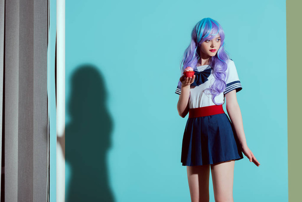 stylish girl in bright wig holding cupcake and looking away in studio - 写真・画像