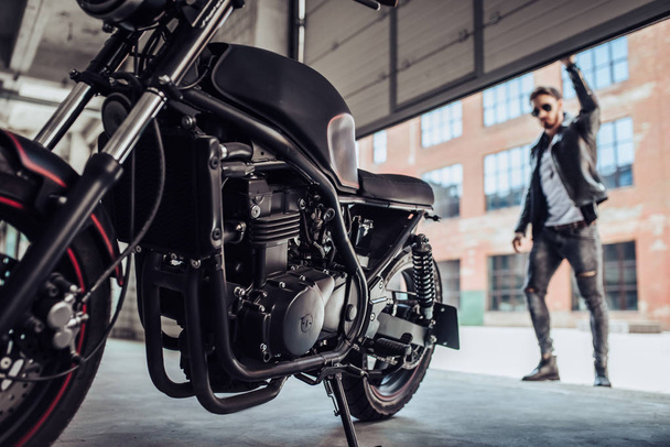 Handsome bearded biker with classic style black motorcycle. Cafe racer in garage. - Fotografie, Obrázek