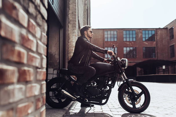 Handsome bearded biker with classic style black motorcycle. Cafe racer in garage. - 写真・画像