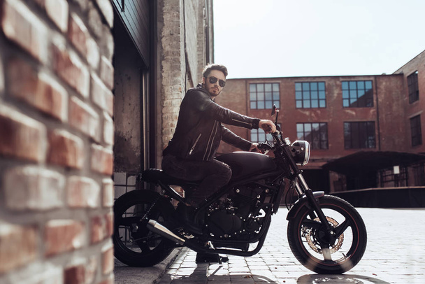 オートバイの古典的なスタイルを黒でハンサムなひげを生やしたバイクに乗る人。ガレージのカフェ レーサー. - 写真・画像