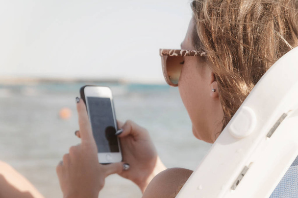 Młoda kobieta opalonej wygląda do smartfona przez okulary przeciwsłoneczne na leżaku. Pojęcie stylu życia jest zawsze w Internecie. - Zdjęcie, obraz