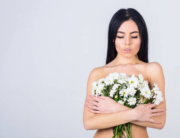 Girl on calm face stands naked and holds chamomile flowers in front of chest. Lady covers breasts with flowers, isolated on grey. Woman with smooth healthy skin looks attractive. Skin care concept - Photo, image