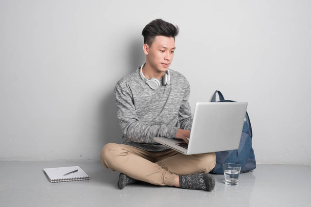 Молодий чоловік сидить на сірій підлозі і використовує ноутбук
. - Фото, зображення