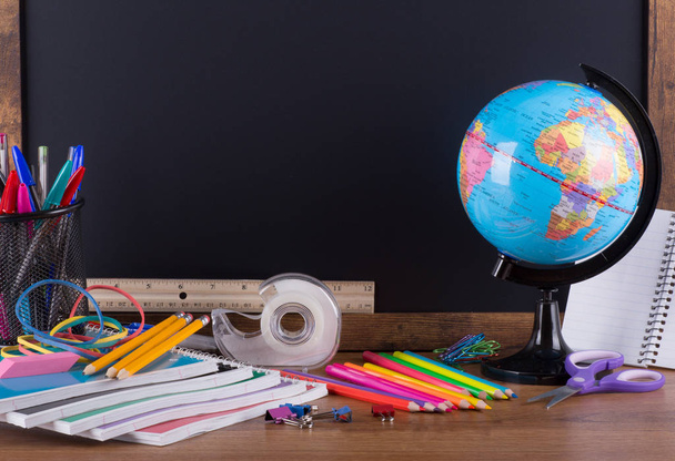 Assortment of back to school supplies on a wooden desktop with blank chalkboard in background for copy space - Foto, Imagem