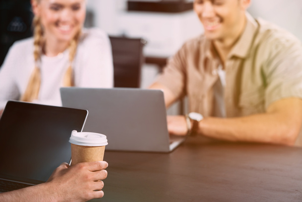 plan recadré d'un homme d'affaires assis à table avec un ordinateur portable et une tasse de café en papier pendant que ses collègues souriants travaillent derrière
  - Photo, image