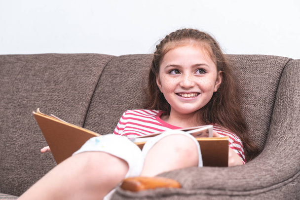 Счастливая маленькая девочка читает книгу на диване
 - Фото, изображение