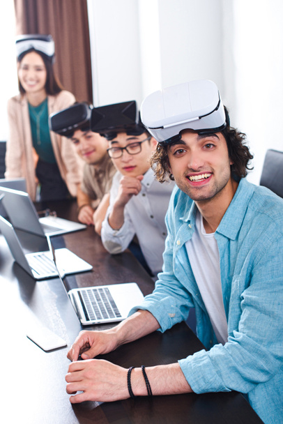groupe souriant de partenaires d'affaires multiethniques avec des écouteurs de réalité virtuelle à table avec des ordinateurs portables dans le bureau moderne
  - Photo, image